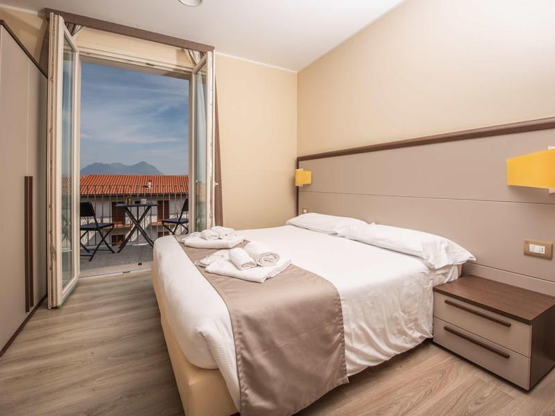 Hell eingerichtetes Hotelzimmer mit Doppelbett, Nachttisch, Balkon und Blick auf rote Dächer.