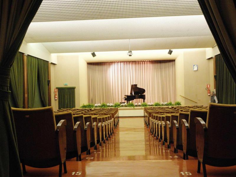 Ein leerer Konzertsaal mit roten Sitzen und einem schwarzen Flügel auf der Bühne vor weißen Vorhängen.