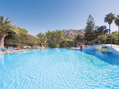 Großer blauer Pool mit Palme, Brücke und Sonnenschirmen vor klarem Himmel und Bergen.