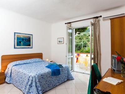 Helles Schlafzimmer mit Holzmöbeln, blau gemustertem Bett, Schreibtisch und offener Terrassentür.