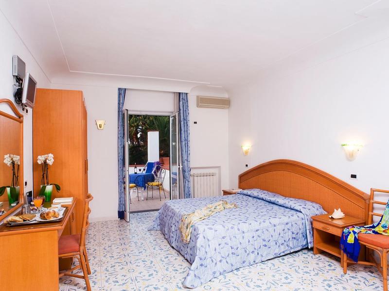 Helles Schlafzimmer mit großem Bett, grünen Möbeln, gelben Vorhängen und Blick auf eine Terrasse.