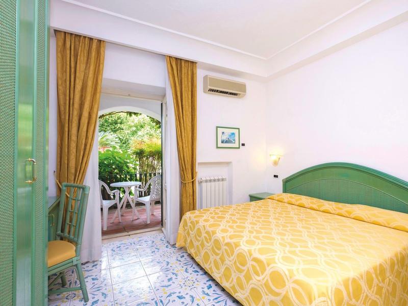 Helles Schlafzimmer mit großem Bett, grünen Möbeln, gelben Vorhängen und Blick auf eine Terrasse.