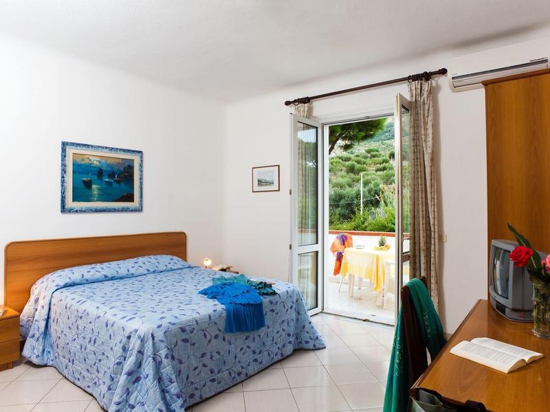 Helles Schlafzimmer mit Holzmöbeln, blau gemustertem Bett, Schreibtisch und offener Terrassentür.