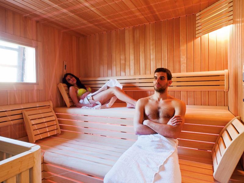 Mann und Frau in einer hellen Holssauna mit mehrlagigen Bänken und Fensterlicht.