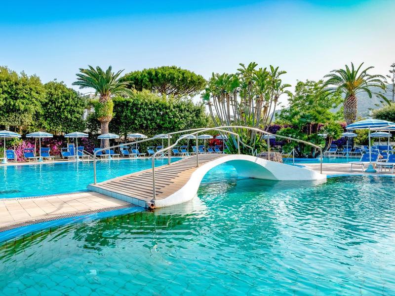 Großer blauer Hotelpool mit weißer Brücke, Palmen und blauen Liegen im Hintergrund.