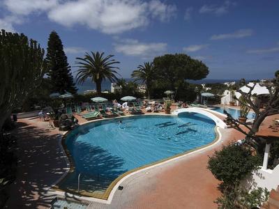 Schwimmbad mit blauem Wasser, umliegenden Liegen, grünen Bäumen und bewölktem Himmel im Hintergrund.