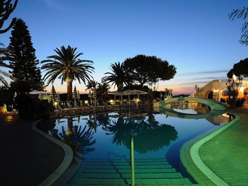 Schwimmbecken bei Sonnenuntergang mit Palmen, blauem Himmel und warm beleuchteten Gebäuden.