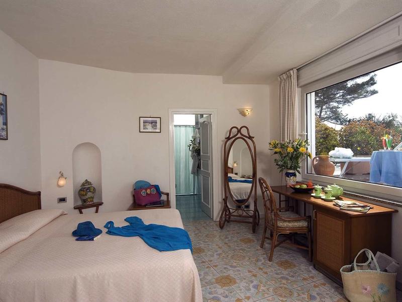 Schlichte Hotelzimmer mit Doppelbett, Schreibtisch, Spiegel, Stuhl und offenem Fenster mit Blick ins Grüne.