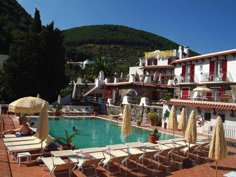 Area piscina con lettini e ombrelloni davanti a un hotel su più piani in un ambiente collinare.