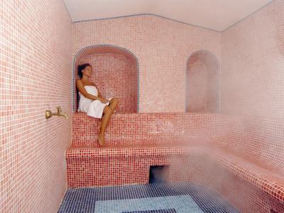 Person relaxing in a steaming pink-tiled steam room with seating and a water basin.