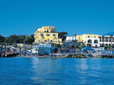 Yellow hotel building next to a white building by the water with boats and blue umbrellas.