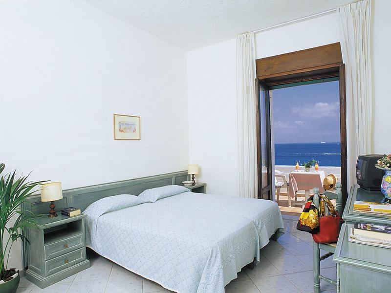 Bright hotel room with double bed, sea view, desk, and balcony with seating area.