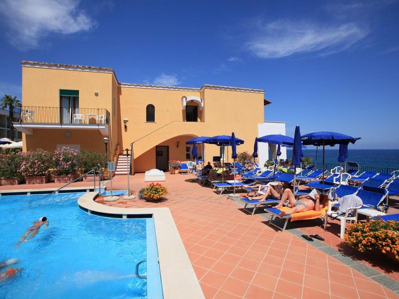 Hotel with pool, sun loungers, and ocean view under a blue sky.