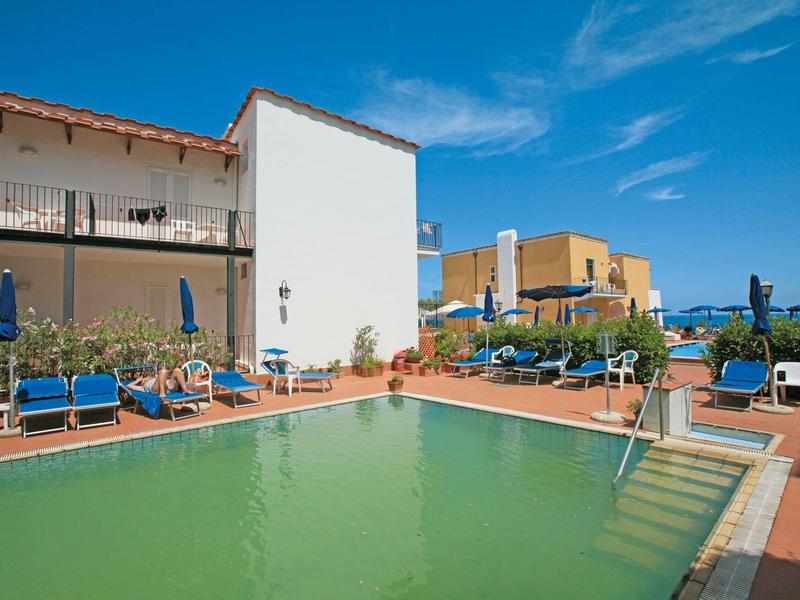 A hotel pool with blue lounge chairs and umbrellas under a clear blue sky.