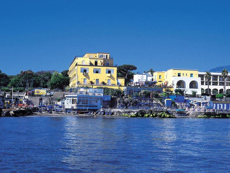 Yellow hotel building by the water under a clear blue sky with surrounding vegetation.