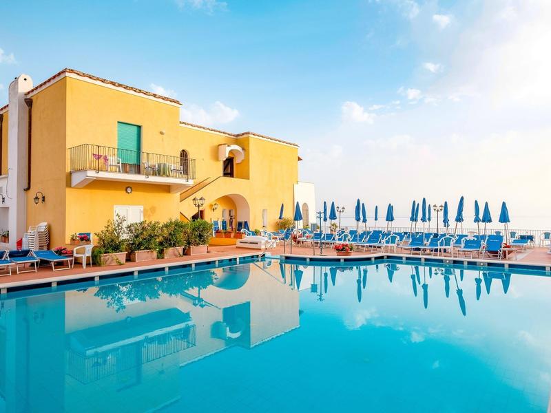 Yellow hotel building next to large swimming pool with blue umbrellas on a sunny day.