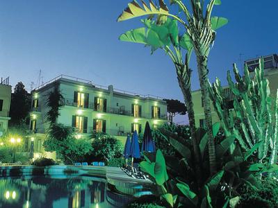 Abendaufnahme eines Hotels mit beleuchtetem Pool und großer Palme im Vordergrund