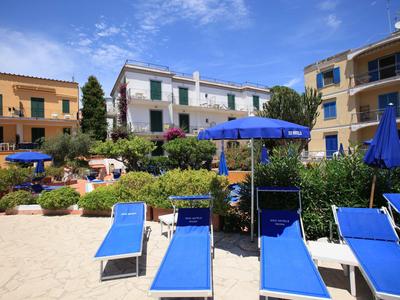 Blick auf blaue Liegestühle und Sonnenschirme vor einem mediterranen Hotel mit Garten unter blauem Himmel.