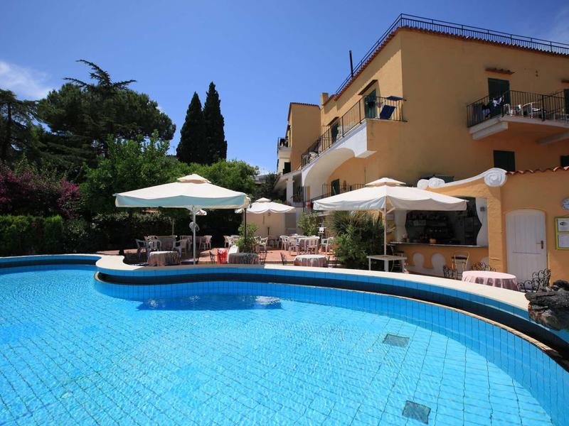 Grande piscine devant un bâtiment d'hôtel jaune avec parasols et jardin