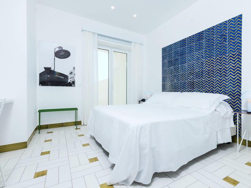 Helles Schlafzimmer mit weißem Bett, blauer Wandgestaltung und Fliesenboden in Weiß und Gelb.