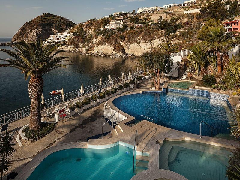 Zwei Schwimmbecken am Meer, umgeben von Palmen und weißen Häusern auf hügeligem Gelände.