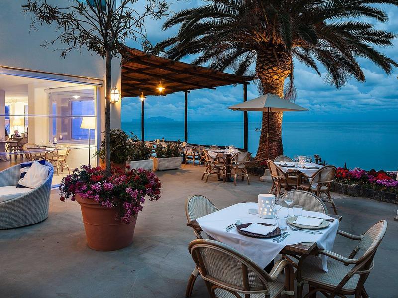 Terrasse mit gedeckten Tischen, Pflanzen und Palmen, Blick auf das Meer bei Abenddämmerung.