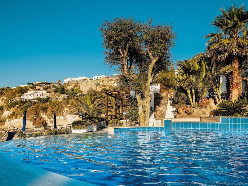 Blauer Pool vor Palmen und Bäumen, im Hintergrund weiße Häuser auf einem Hügel unter blauem Himmel.