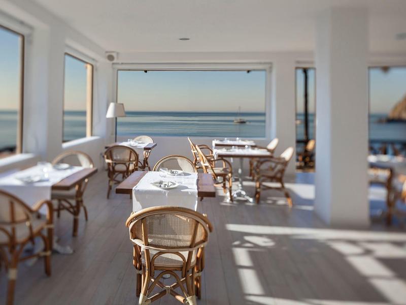 Helles Restaurant mit Holzstühlen, Tischen und Blick auf ruhiges Meer durch große Fenster.