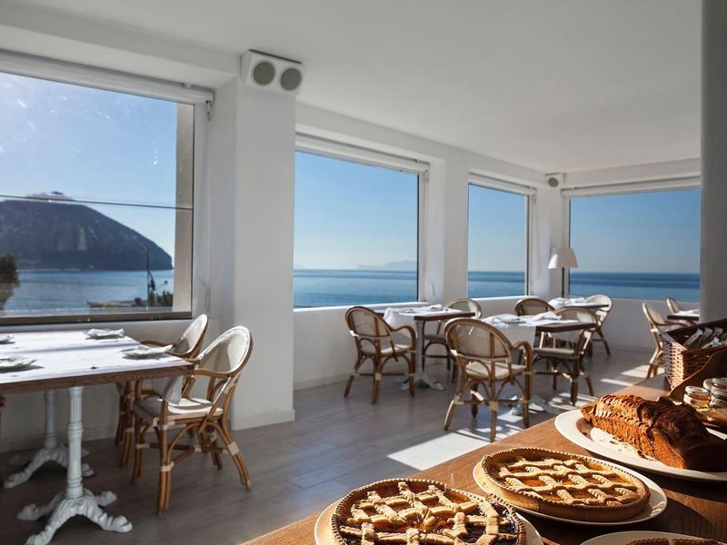 Helles Café mit weißen Wänden, Holzstühlen und Tischen, großer Fensterblick aufs Meer und Gebirge.