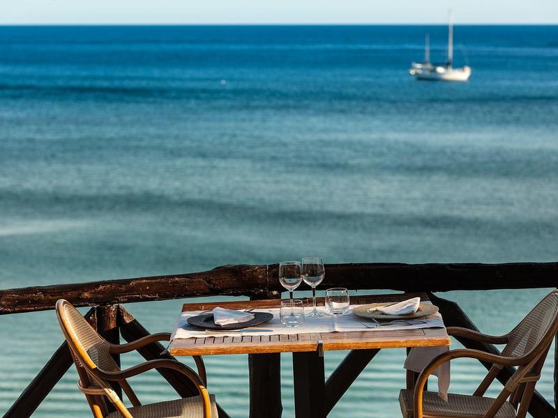 Holztisch mit zwei Stühlen auf Balkon, gedeckt mit Gläsern, Blick auf ruhiges blaues Meer und Segelboot.