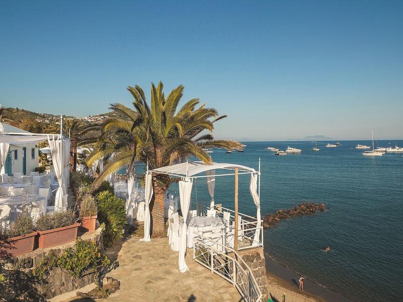 Küste mit Palmen, weißer Terrasse und Booten auf blauem Meer unter klarem Himmel.