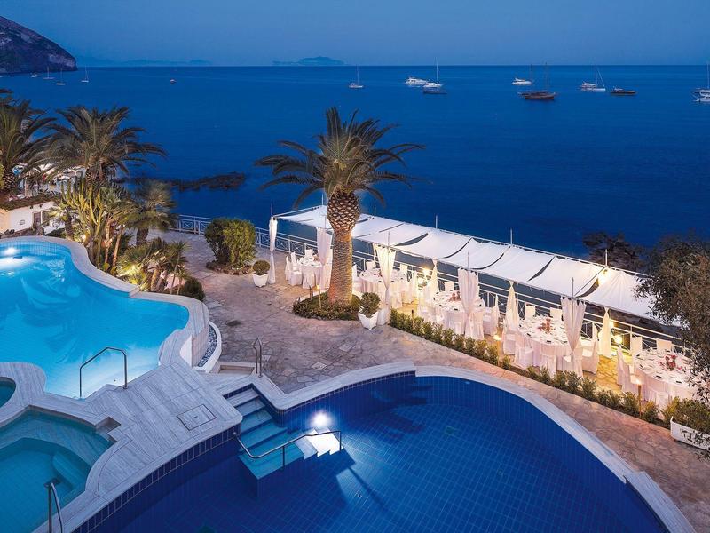 Blick auf zwei ineinander übergehende Pools mit Palmen und Meer mit Booten im Hintergrund.