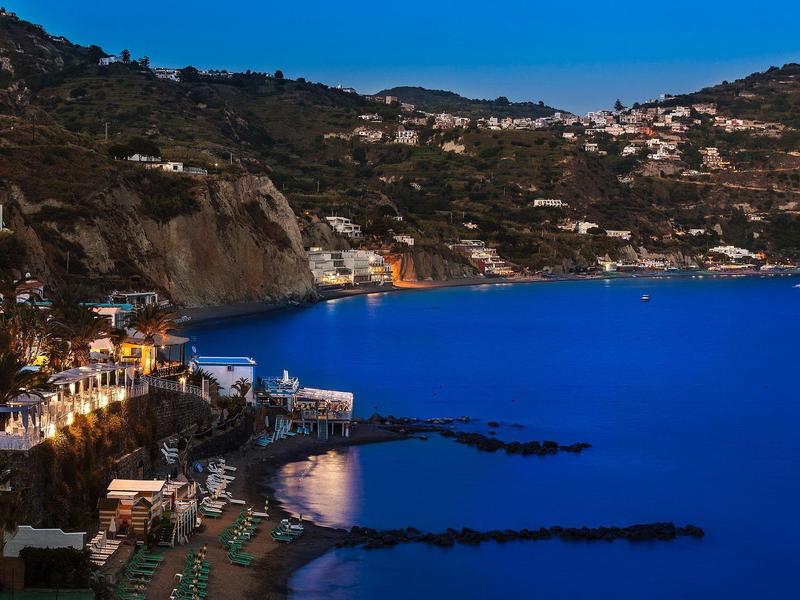 Küstenstadt bei Nacht mit leuchtendem Meer, Hügeln und Häusern, die im Dunkeln erstrahlen.