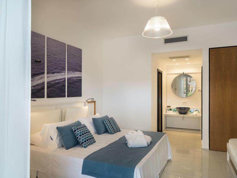 Hell eingerichtetes Schlafzimmer mit Doppelbett, blauen Kissen, Deckenbeleuchtung und offenem Bad.