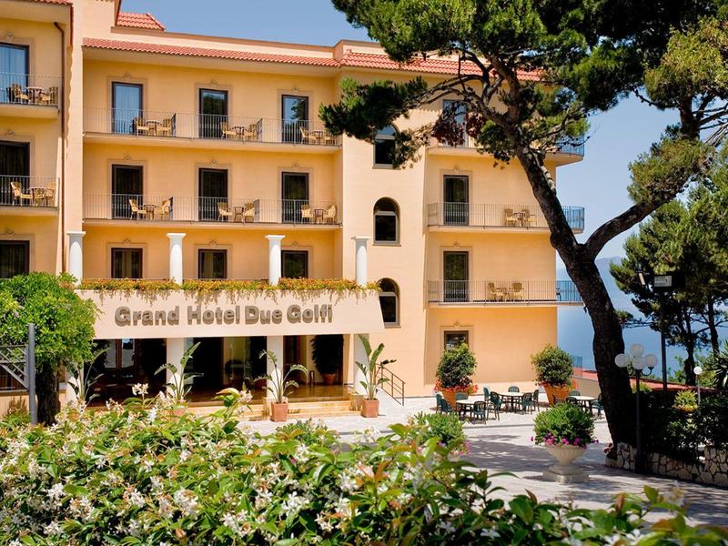 Gelbes Hotelgebäude mit Balkonen, grünen Pflanzen und blauen Himmel am Meer im Hintergrund.