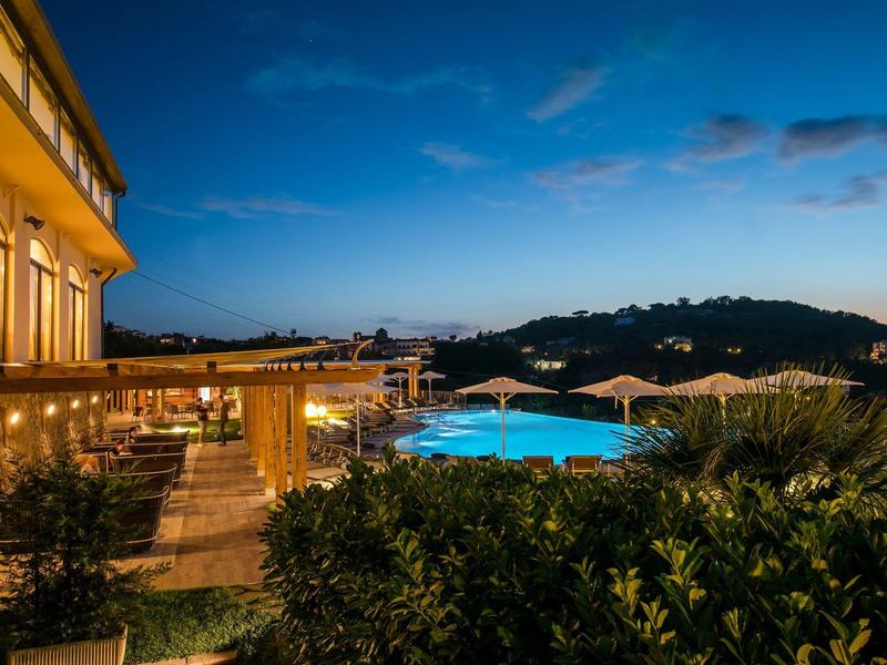 Abendblick auf einen beleuchteten Hotelpool und Terrassen mit Hügeln im Hintergrund