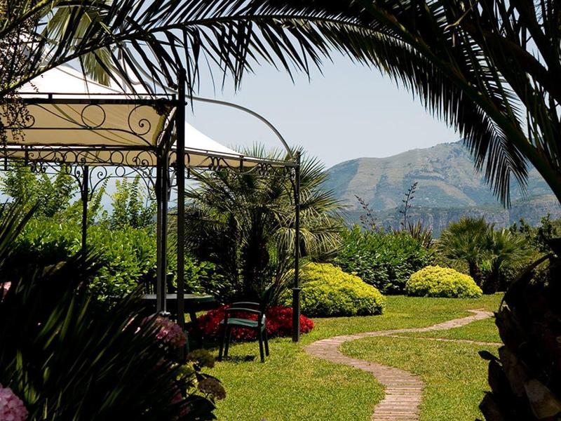 Grüner Garten mit bunten Blumen, Palmen und einem Weg vor Bergen im Hintergrund bei klarem Himmel.