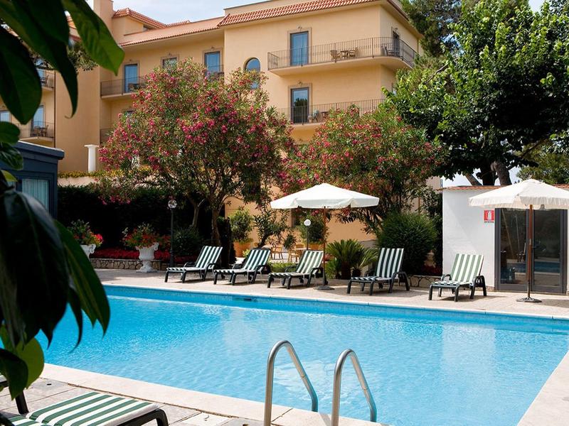 Hotelpool mit Liegestühlen und Sonnenschirmen vor einem gelben Gebäude mit zwei Stockwerken