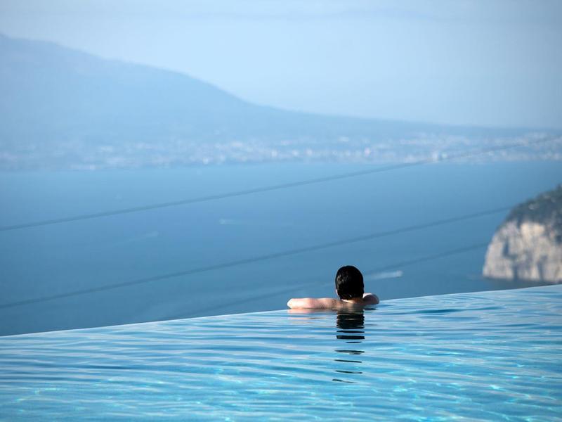 Person entspannt sich im Infinity-Pool mit Blick auf Berge und Küste.