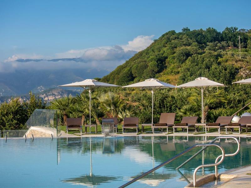 Moderner Pool mit Liegestühlen und Sonnenschirmen vor grünen Hügeln und blauem Himmel.