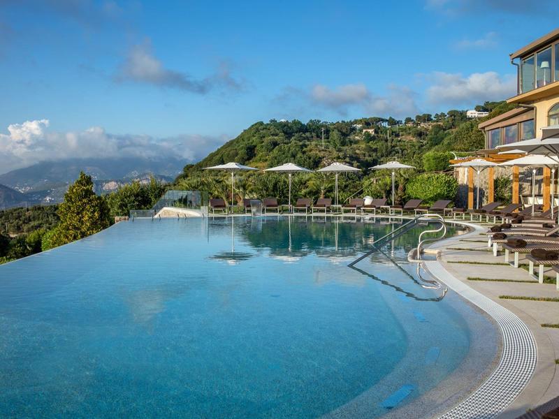 Infinity-Pool mit Liegestühlen und Sonnenschirmen vor hügeliger Landschaft und blauem Himmel.