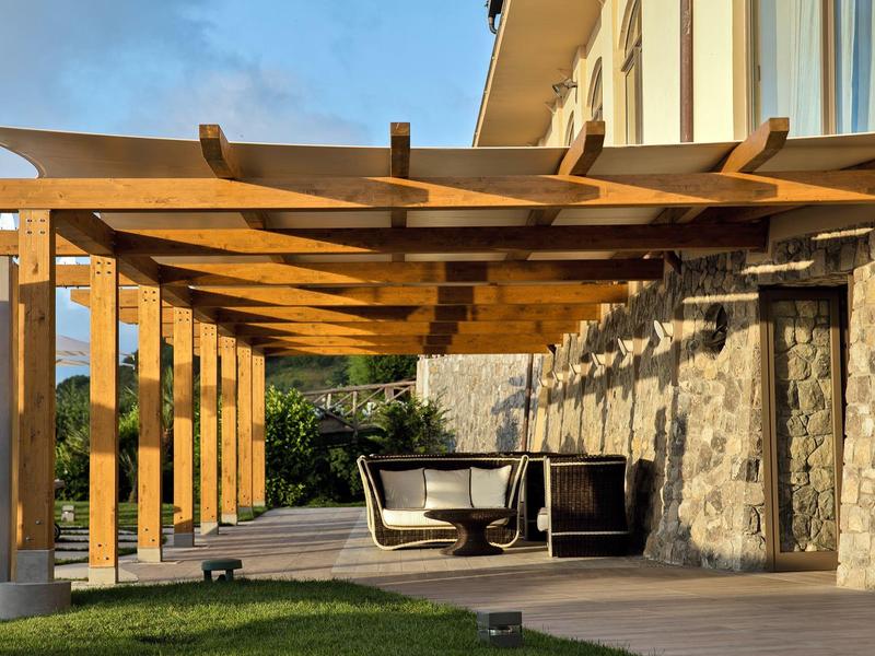 Holzpergola über einer Terrasse mit Gartenmöbeln und Steinmauer im Hintergrund.