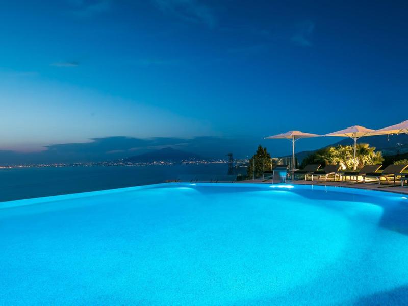 Beleuchteter Infinity-Pool mit Meerblick und Lounge-Bereich bei Dämmerung.