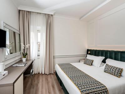 Hell eingerichtetes Hotelzimmer mit Doppelbett, dunklem Samtkopfteil und Fenster mit Gardinen.