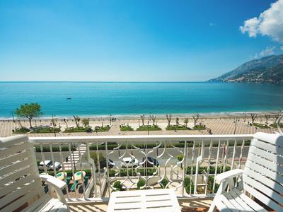 Blick von einem Balkon mit weißen Stühlen auf einen Sandstrand und blaues Meer unter klarem Himmel.