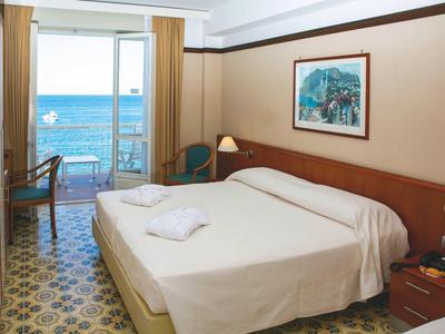 Hotelkamer met tweepersoonsbed, balkon met uitzicht op zee, lichte inrichting en patroonvloer.