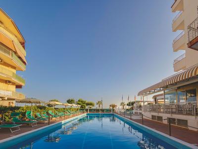 Schwimmbad zwischen Hotelgebäuden mit Sonnenliegen und Sonnenschirmen unter klarem blauem Himmel.
