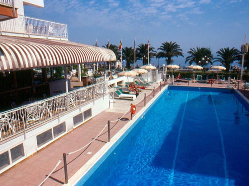 Groot buitenzwembad met ligstoelen en parasols naast een hotelterras met luifel.