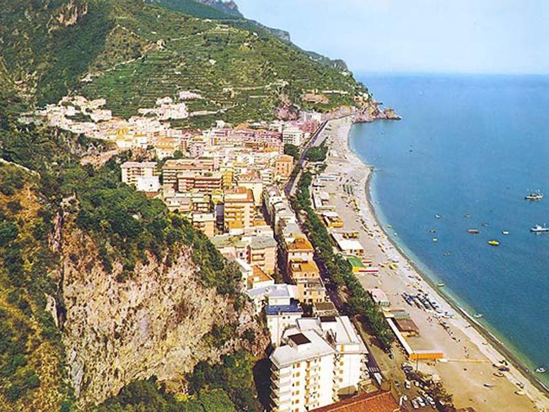 Luchtfoto van een kustplaats met strand, gebouwen en beboste heuvels.