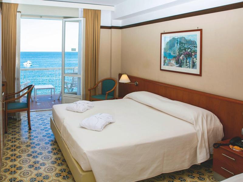 Hotelkamer met tweepersoonsbed, balkon met uitzicht op zee, lichte inrichting en patroonvloer.